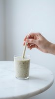 Hand holding reusable straw in creamy smoothie drink with seeds and herbs in glass on white marble table for healthy beverage and sustainable lifestyle content.