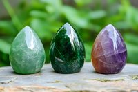 Three polished gemstone eggs in green aventurine, malachite and amethyst arranged on wooden surface with natural foliage background for crystal healing meditation.