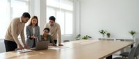 Diverse business team collaborating around laptop in bright modern conference room with natural lighting and minimalist design for corporate meetings.