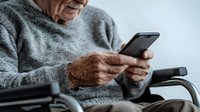 Senior Caucasian man in gray sweater sitting in wheelchair using smartphone, cropped view focusing on hands holding mobile device. Modern elderly lifestyle.