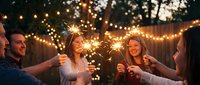 Group of young adults holding sparklers at outdoor evening celebration with warm string lights and trees in background, suitable for party, event, and lifestyle content.