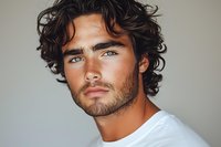 Young Hispanic man with curly dark hair and defined facial features wearing white t-shirt against neutral background, looking intensely at camera. Professional headshot style portrait.