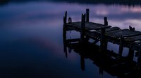 Abandoned wooden pier extending into calm water at twilight with purple and blue sky reflecting on still surface.
