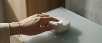 Woman hands gently touching white geometric cube on minimalist table surface in soft natural lighting for modern interior design concepts.