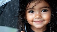 Young Asian girl with bright smile holding black umbrella in rain, close up portrait with natural lighting and bokeh raindrops creating magical atmosphere.