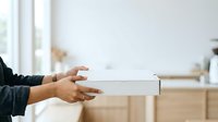 Person in casual black long-sleeve shirt holding white cardboard box with both hands in bright modern interior space, suitable for delivery, shipping, moving, and e-commerce concepts.