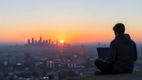 Young professional man working on laptop computer overlooking city skyline at golden hour sunset. Remote work and digital nomad lifestyle concept.