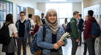 Young Middle Eastern female student wearing hijab smiling in busy school hallway holding books and backpack during break time between classes.