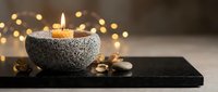 Spa candle burning in stone bowl on dark slate with warm bokeh lights creating peaceful wellness atmosphere for relaxation and meditation rituals.