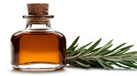 Small glass bottle with cork stopper containing amber liquid next to fresh rosemary sprig on white background, suitable for aromatherapy and wellness concepts.