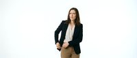 Woman with long dark hair and olive complexion wearing professional business attire including black blazer and beige trousers, standing confidently against white background for corporate portraits.