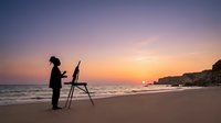 Silhouette of woman artist painting on easel at sunset beach with golden sky and ocean waves, creative outdoor workspace.
