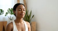 Peaceful young African American woman meditating with closed eyes in modern home interior with green plants, practicing mindfulness and wellness routine.