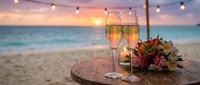 Romantic beach sunset celebration with champagne glasses and tropical flowers on wooden table with string lights creating perfect atmosphere for wedding or anniversary.
