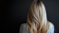 Blonde woman with long straight layered hair shown from behind against dark background, demonstrating modern hairstyling and highlighting techniques.