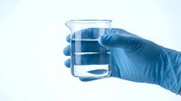Hand in blue medical glove holding glass beaker with clear liquid for laboratory research and scientific testing on white background.