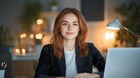 Young Caucasian businesswoman with red hair wearing black blazer and white shirt smiling while working at laptop in cozy home office with warm lighting.