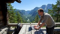 Young Caucasian man working on laptop at mountain cabin terrace with dramatic alpine valley landscape and forest views during summer vacation remote work.