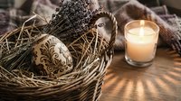 Rustic Easter composition with decorated wooden egg in wicker basket, dried lavender bouquet and glowing candle on textured wooden surface for spring decor.