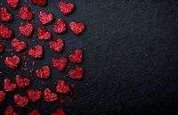 Red sugar heart-shaped sprinkles scattered on dark slate background with copy space, romantic food styling for Valentine's Day celebrations.