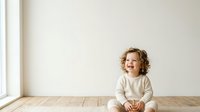 Young child with curly blonde hair, appearing to be a toddler of Caucasian ethnicity, wearing cream-colored clothing, sitting on wooden floor in bright, minimalist interior space with white walls and natural light from window.