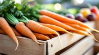 Fresh organic carrots with green tops in wooden crate at farmers market with colorful vegetables in background for healthy cooking and nutrition.