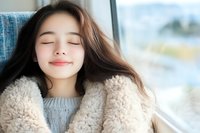 Young Asian woman with closed eyes and gentle smile wearing cozy gray sweater and fluffy beige coat, expressing serenity and contentment near window with natural light.