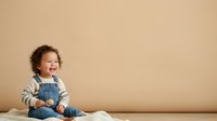 Young toddler with curly brown hair and fair complexion wearing denim overalls and striped long-sleeve shirt, sitting against neutral beige background. Suitable for parenting, childcare, and family lifestyle content.