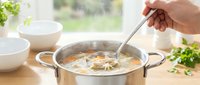 Hand stirring homemade vegetable soup in stainless steel pot on kitchen counter. Fresh herbs and bowls nearby create cozy cooking atmosphere for recipes.