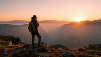 Female hiker with backpack standing on mountain peak at sunrise, overlooking misty valleys and distant mountains during golden hour outdoor adventure.