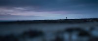 Solitary lighthouse standing on distant horizon at dusk, moody twilight seascape with dramatic cloudy sky and calm waters for atmospheric coastal backgrounds.