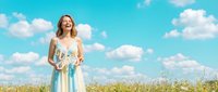 Young woman with blonde hair, fair complexion, wearing light blue dress standing in wildflower meadow under bright sky. Suitable for lifestyle, wellness, freedom and nature themes.