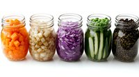 Glass jars with colorful pickled and fermented vegetables including carrots, onions, red cabbage, cucumbers and peppercorns arranged in row on white background.