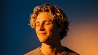 Young Caucasian man with curly hair meditating peacefully at sunset with closed eyes and serene expression against dramatic orange and blue sky backdrop.