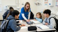 Female teacher with glasses instructing diverse elementary students at classroom table during group learning activity in modern educational setting.