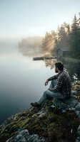 Bearded man in plaid shirt sitting on rocky shore contemplating misty lake at sunrise with wooden dock and cabin surrounded by evergreen forest.