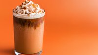 Iced chocolate frappe topped with whipped cream and cocoa powder in tall glass against vibrant orange background, cold refreshing drink with condensation drops.