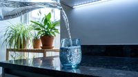 Fresh clean water pouring from modern faucet into glass on kitchen counter with green houseplants creating healthy hydration lifestyle scene.
