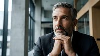 Thoughtful mature Caucasian businessman with gray beard in dark suit sitting in modern office, hands clasped under chin in contemplative pose.