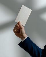 Male hand in business suit holding up blank white card against gray wall with shadow overlay, mockup for business card or corporate identity design.