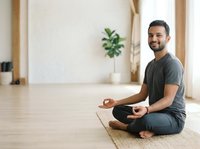 Young Middle Eastern man meditating in lotus position on yoga mat in bright studio space with wooden floors and potted plant, practicing mindfulness and wellness.