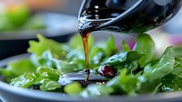 Pouring dark balsamic vinegar dressing over fresh green salad leaves creates glossy stream in close-up view, highlighting culinary preparation and healthy eating.