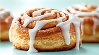 Fresh baked cinnamon roll with sweet white vanilla icing dripping down sides, closeup view of spiral pastry showing flaky layers on mint green background.
