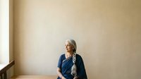 Mature woman with gray braided hair wearing dark blue shirt standing against neutral beige wall in minimalist interior space for portrait and lifestyle use.