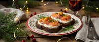 Festive holiday appetizers with smoked salmon on dark bread garnished with herbs and cream cheese, served on decorative plate with Christmas lights background.