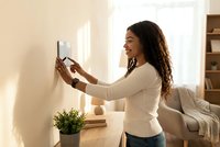 Young Hispanic woman adjusting smart home thermostat on wall in modern living room with natural sunlight for energy efficiency and comfort control.