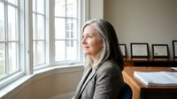 Senior woman with long gray hair and light skin tone, wearing a beige blazer, sitting at wooden desk by large windows in bright office space, looking thoughtfully out window.