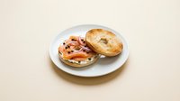 Toasted bagel with smoked salmon, cream cheese, capers, and red onion on a white plate against a beige background.