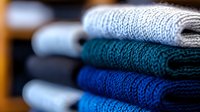 Stack of neatly folded warm knitted sweaters in various shades of blue and gray, showing detailed wool texture and cozy winter clothing arrangement.