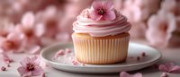 Delicate vanilla cupcake with pink frosting swirl decorated with cherry blossom flowers and petals on white plate against soft floral background.